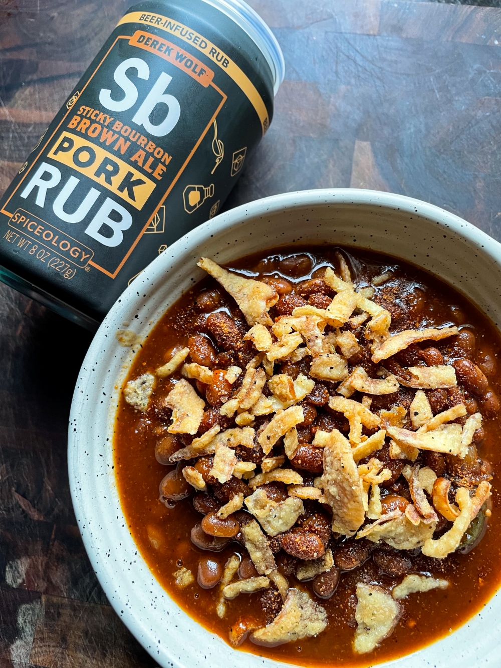 Bowl of Smoked BBQ Beans topped with crispy onions next to a can of Spiceology X Derek Wolf Sticky Bourbon Brown Ale beer-infused rub